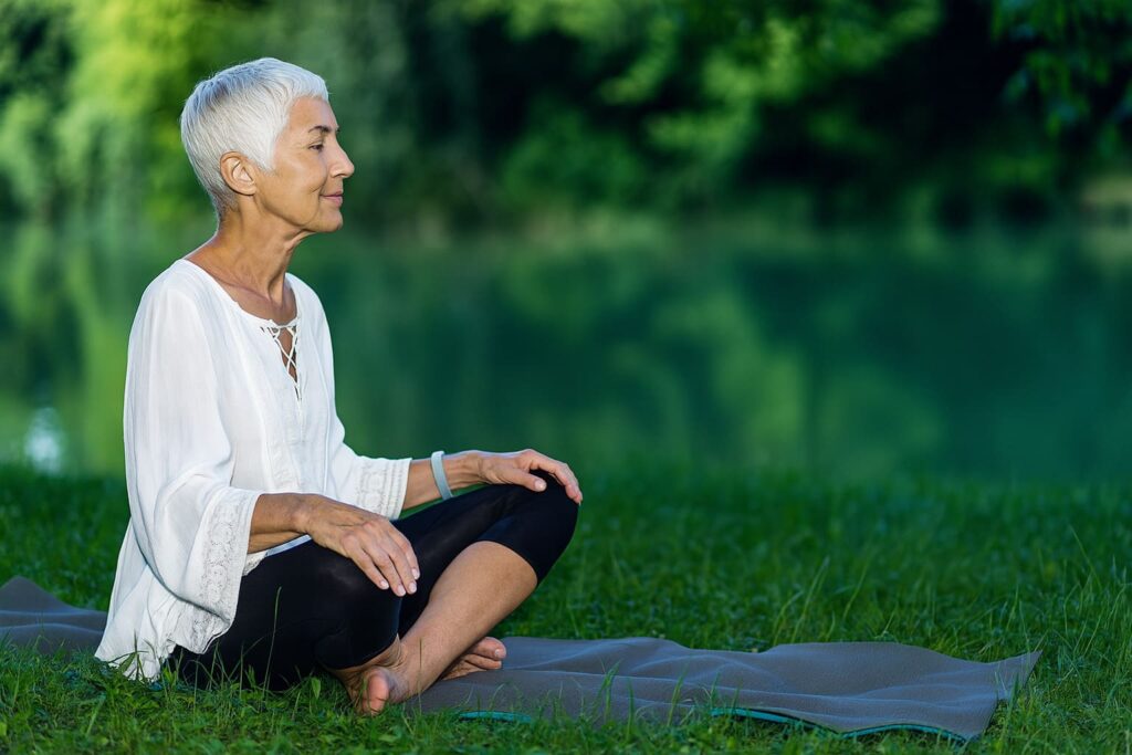 Starsza kobieta medytująca na macie do jogi na tle spokojnego jeziora i zieleni natury.